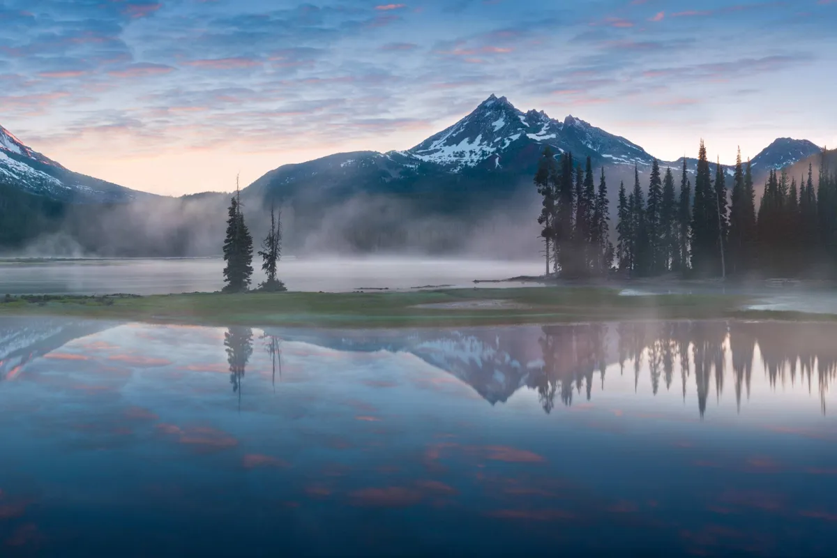 A panoramic view showcasing a tranquil mountain lake surrounded by dense trees and a dramatic sky, with distant peaks adding to the wilderness feel. The natural setting is ideal for hiking, wildlife spotting, and capturing stunning sunrises or sunsets.