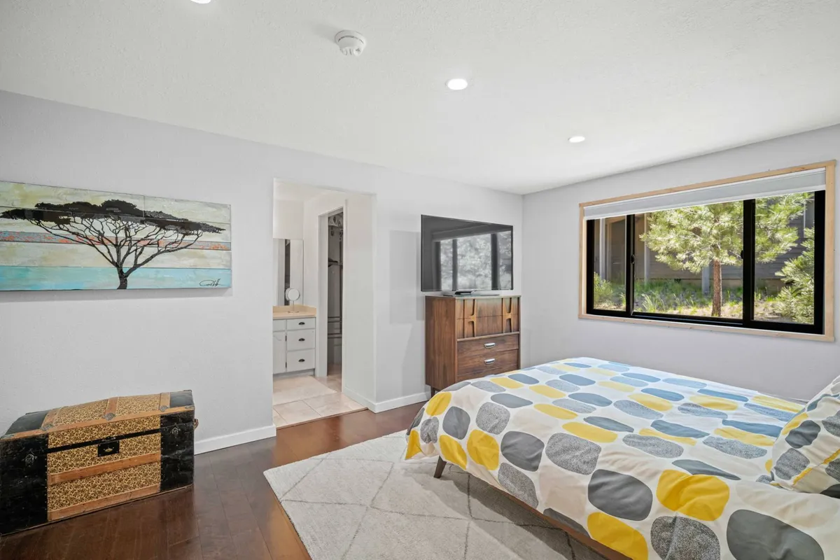 A serene bedroom with a bed adorned with a patterned duvet, a large window offering views of greenery, wall artwork, and a wooden dresser with a TV. The mood is peaceful and restorative, perfect for unwinding.