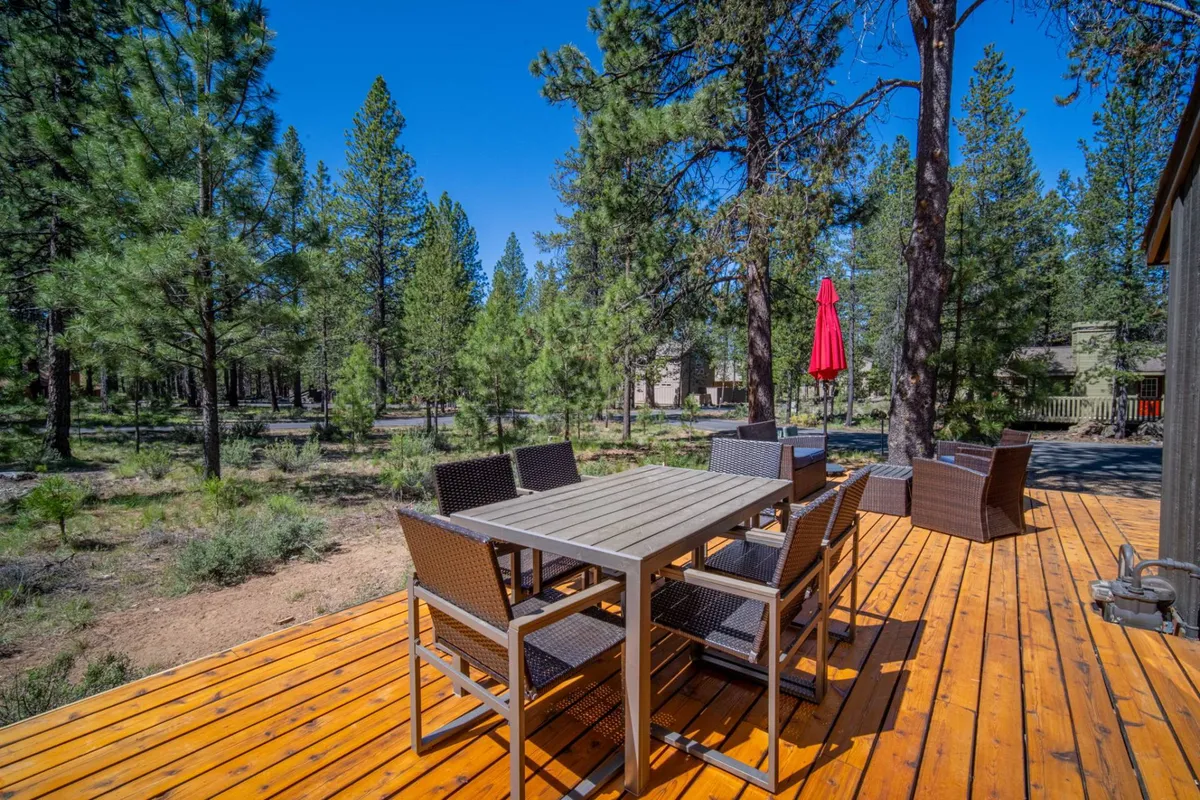 A spacious wooden deck nestled among trees features an outdoor dining table, chairs, and a vibrant red umbrella. The setting invites gatherings, meals, and leisure in a relaxed, nature-surrounded atmosphere.