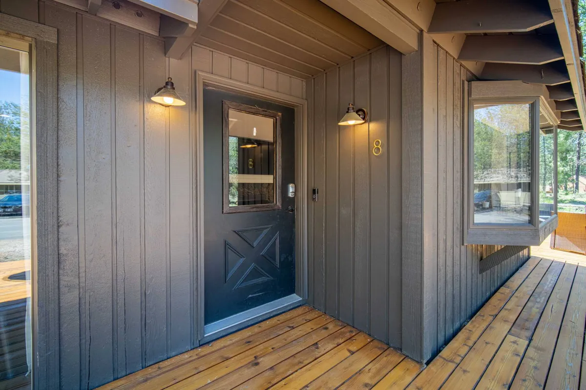 A welcoming view of the home's front entrance featuring a wooden deck, warmly lit front door, and surrounding trees. The scene suggests a hospitable return after a day spent exploring snowy trails or hitting the slopes.