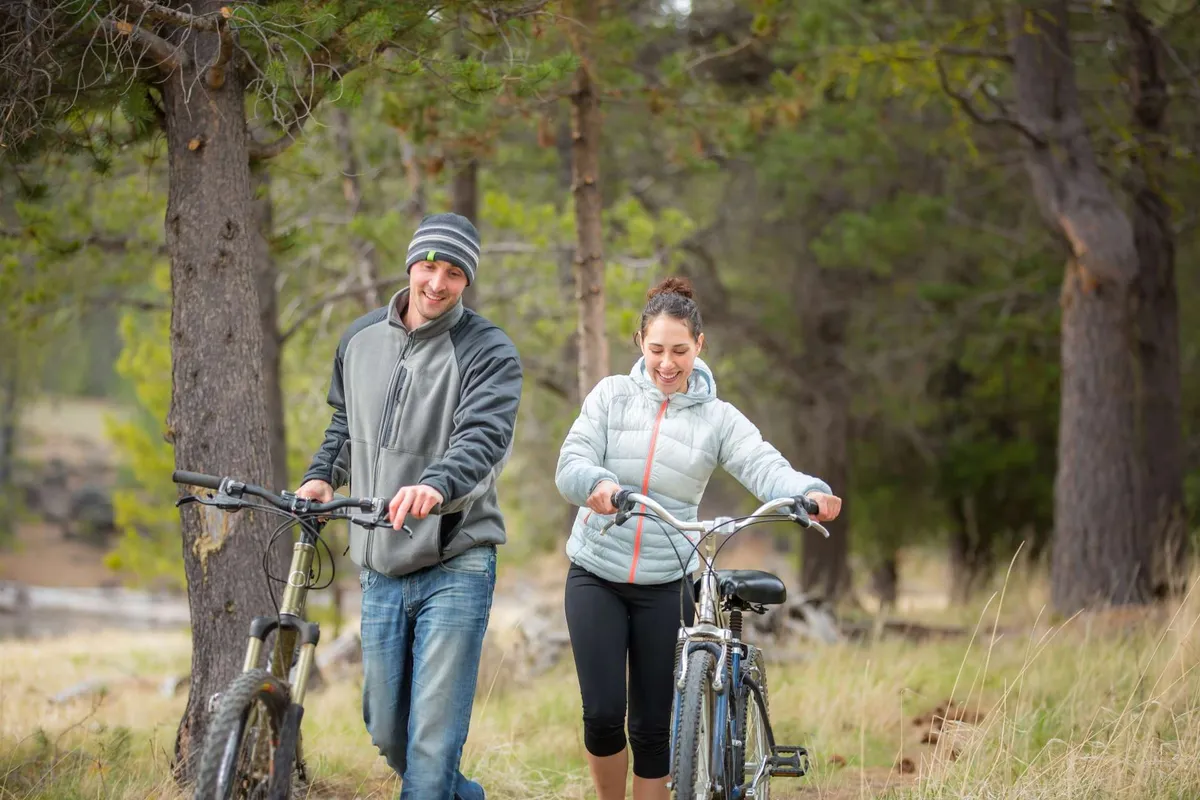 This exterior image features two cyclists walking with bicycles amidst a lush green landscape and tall trees, capturing the spirit of outdoor adventure and Sunriver’s famed paved trails. The relaxed, joyful mood highlights the biking opportunities available directly from the property.