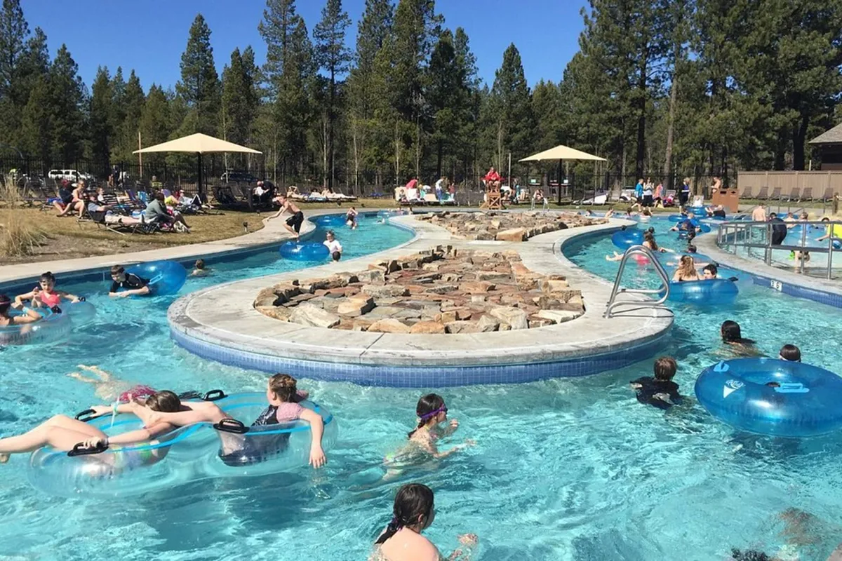 A lively exterior shot of a resort-style swimming pool, complete with rock formations, people floating on inner tubes, and umbrellas amid tall pine trees. The joyful, fun mood perfectly illustrates the kind of family-friendly recreation offered at SHARC, which is included in the stay.
