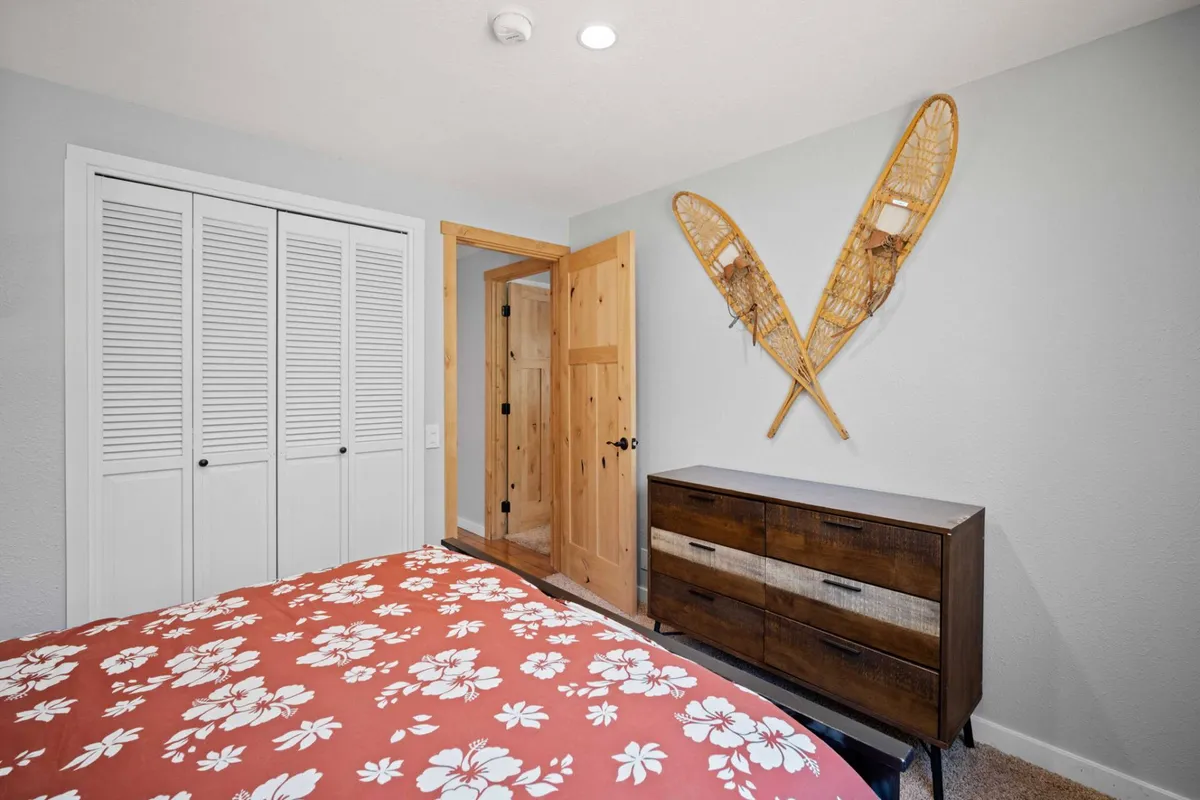 This image features a bed with a floral pattern, a wooden dresser, and decorative snowshoes on the wall. The space is bright and cozy, with soft lighting and cabin-inspired decor that evokes warmth and relaxation.