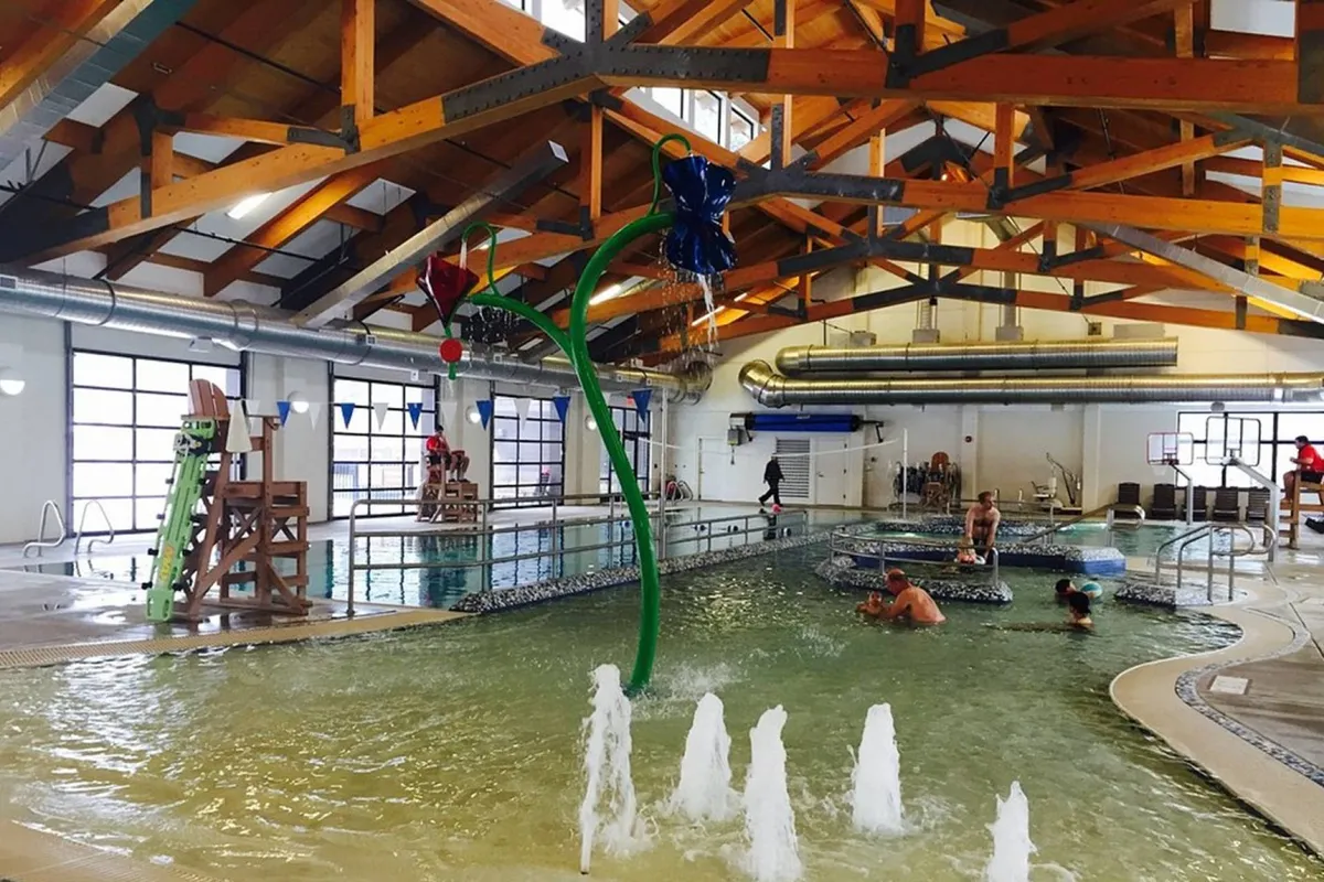 A lively interior recreation space filled with water play structures, swimming pools, and families enjoying themselves under wooden beams. Children and adults alike are engaged in swimming, splashing, and socializing, capturing the communal spirit of Sunriver Resort.
