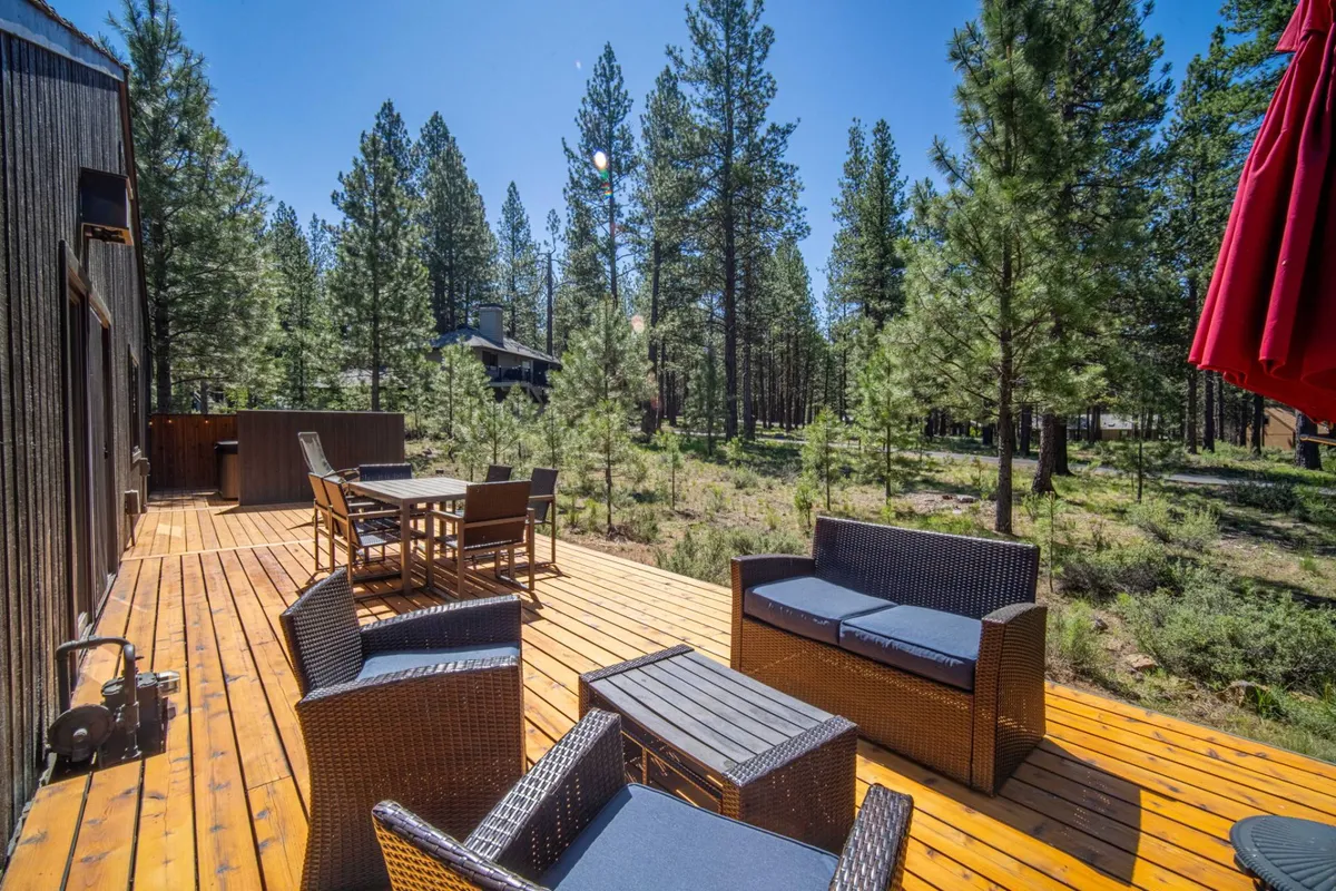 The image captures an open wooden deck with ample outdoor seating surrounded by towering pine trees. The scene radiates tranquility and natural beauty, evoking moments of relaxing, socializing, and soaking up sunset views through the trees.