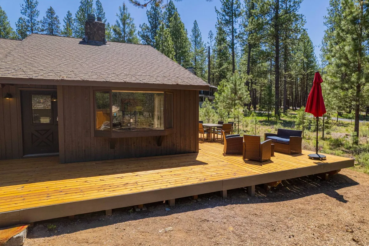 A tranquil exterior view showcasing a spacious wooden deck with outdoor furniture and a red umbrella, surrounded by lush green trees. The inviting seating area hints at leisurely outdoor meals, nature-watching, and socializing in the peaceful forest setting.