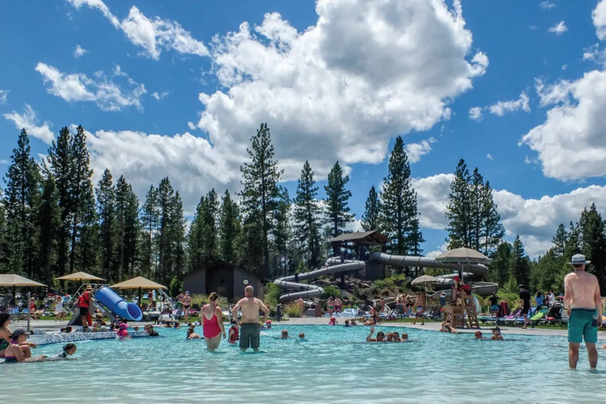 An exterior view of a vibrant swimming pool with a water slide, surrounded by green trees. Families and children are splashing, swimming, and socializing, infusing the space with energy and joy.