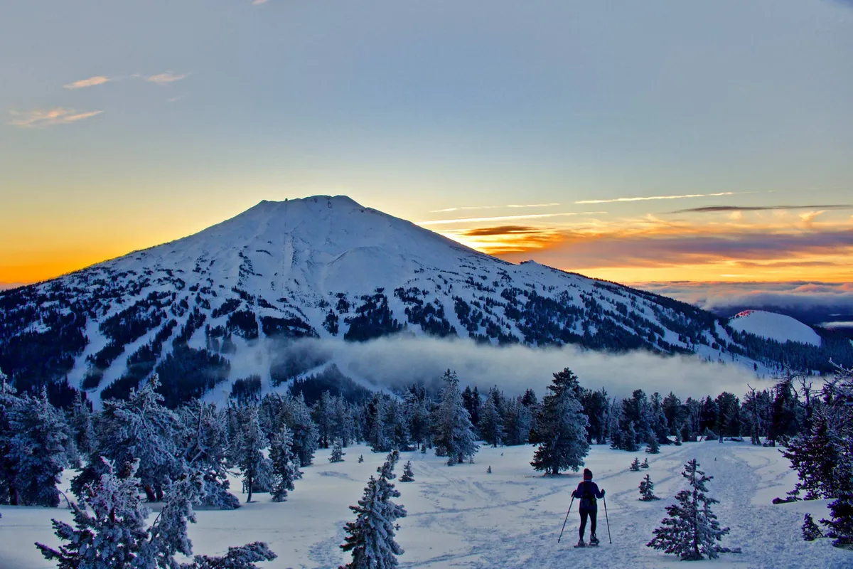 A breathtaking view of a snow-covered mountain at sunset, with a skier in the foreground gliding across the pristine slopes. Evergreen trees dot the scene, capturing the adventurous spirit of winter sports and the tranquil beauty of the mountain landscape.
