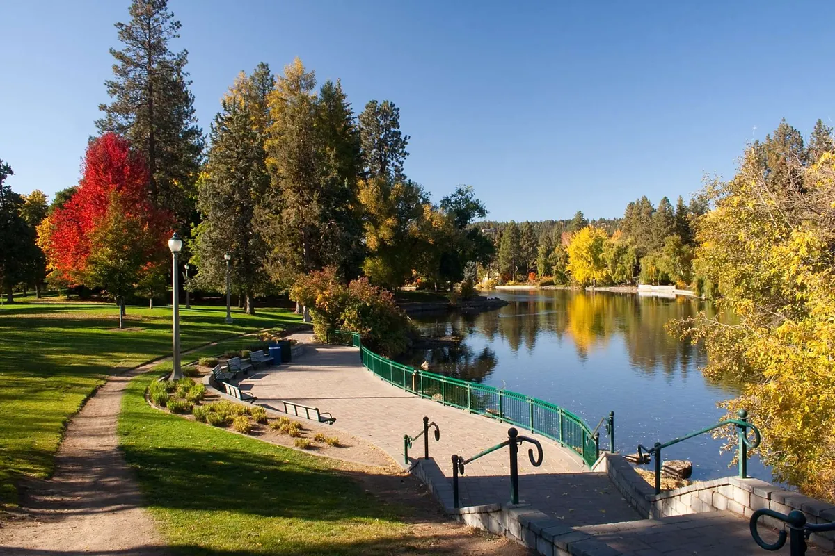 A tranquil lakeside walkway winds past colorful trees and calm water, with park benches and a charming bridge in view. The scene exudes peace and provides a glimpse of the area’s natural beauty, ideal for biking, walking, or spotting wildlife.