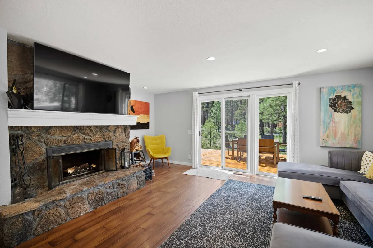 A cozy living room scene featuring a prominent wood-burning fireplace, a large TV, a cheerful yellow chair, and sliding doors leading to the deck. Artwork and natural views add warmth, while the glass sliders suggest an easy transition to outdoor spaces.